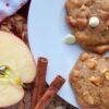 Two applesauce cinnamon cookies with white chocolate chips on a white plate, near a halved apple and cinnamon sticks.