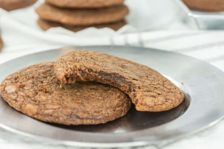 Two chocolate cookies are placed on a metal plate, with the cookie on top partially eaten. In the background, a blurred stack showcases more of these delicious treats made from an easy 3 Ingredient Nutella cookies recipe.