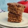 Two stacked square apple cider blondies on a white plate, with an out-of-focus red apple and folded plaid cloth in the background.