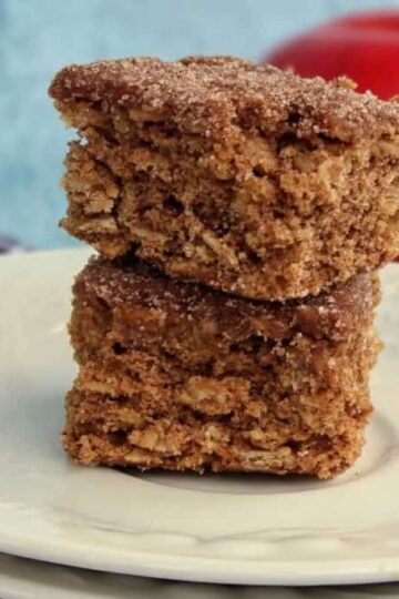 Two stacked square apple cider blondies on a white plate, with an out-of-focus red apple and folded plaid cloth in the background.