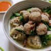 A bowl of Indonesian meatballs in soup with celery, a bowl or chili sauce, and a bowl of fried shallots nearby.