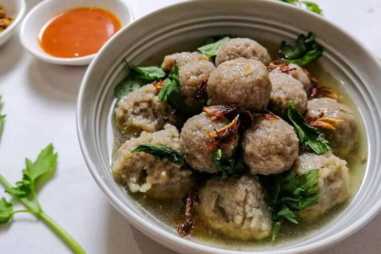A bowl of Indonesian meatballs in soup with celery, a bowl or chili sauce, and a bowl of fried shallots nearby.