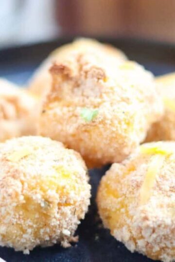 A plate with six breaded and baked cheesy potato balls, garnished with green herbs, next to a fork.