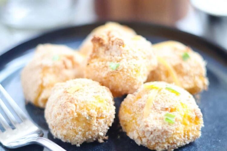 A plate with six breaded and baked cheesy potato balls, garnished with green herbs, next to a fork.