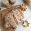 A decorative bread shaped like a Christmas tree on a wooden board, with a yellow star-shaped cutter, two spice containers, and a painted wooden spoon on a burlap mat.