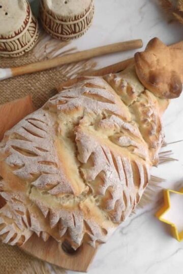 A decorative bread shaped like a Christmas tree on a wooden board, with a yellow star-shaped cutter, two spice containers, and a painted wooden spoon on a burlap mat.