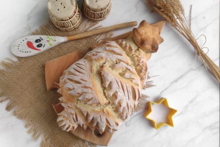 A decorative bread shaped like a Christmas tree on a wooden board, with a yellow star-shaped cutter, two spice containers, and a painted wooden spoon on a burlap mat.