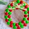 A doughnut decorated with red and green candies and white icing, arranged to resemble a holiday wreath, is placed on a white textured cloth.