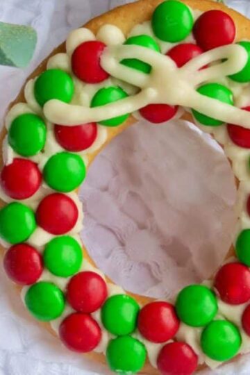 A doughnut decorated with red and green candies and white icing, arranged to resemble a holiday wreath, is placed on a white textured cloth.