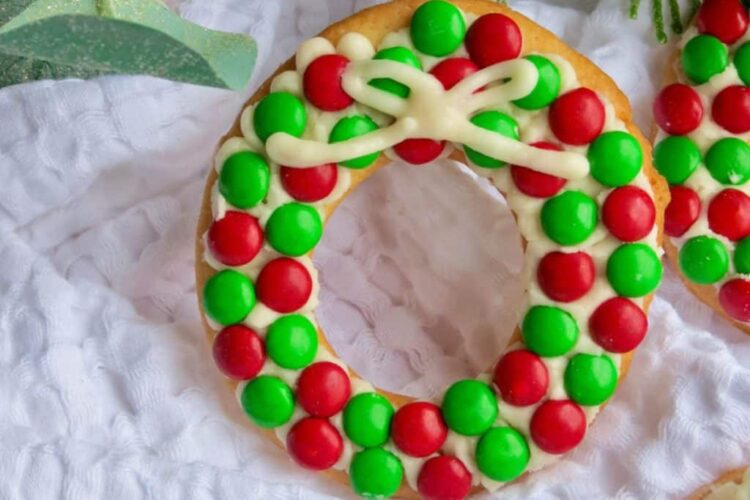 A doughnut decorated with red and green candies and white icing, arranged to resemble a holiday wreath, is placed on a white textured cloth.