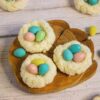 Coconut macaroons shaped like nests, each holding colorful candy eggs, create a charming display on a wooden board and white surface. This delightful coconut Easter egg recipe for cookies adds a festive touch to any gathering.