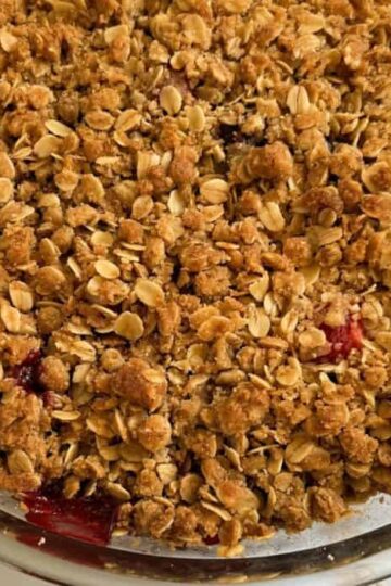 A round, glass dish contains a delightful strawberry crisp with oatmeal, featuring a golden-brown oat topping and edges revealing a luscious blackberry and red fruit filling.