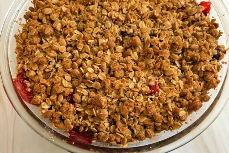 A round, glass dish contains a delightful strawberry crisp with oatmeal, featuring a golden-brown oat topping and edges revealing a luscious blackberry and red fruit filling.