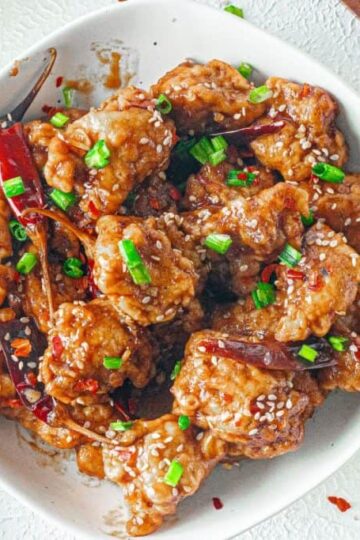 A bowl of crispy General Tso chicken pieces garnished with sesame seeds, chopped green onions, dried red chilies, and served with rice and broccoli on the side.