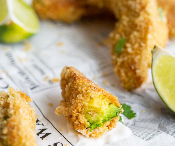 A side view showing the bite taken out of an avocado fry on a newspaper with limes in the background.