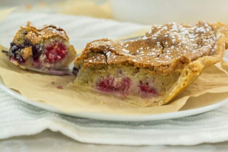 A slice of fruit-filled pie with a golden crust and powdered sugar sits on a plate, next to a partially eaten piece on a fork.