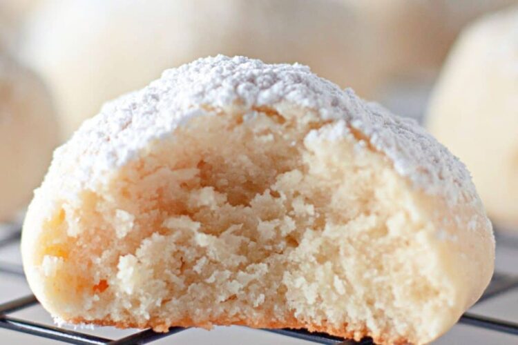 A close-up of a powdered sugar-coated cream cheese cookie with a bite taken out, resting on a wire cooling rack.