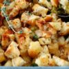 Close-up of bread stuffing with herbs in a blue baking dish. A sprig of thyme is placed on top, and a spoon is visible on the side.