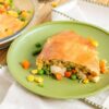 A slice of turkey pot pie on a green plate, filled with chicken, peas, carrots, corn, and a golden-brown crust, with a pie dish in the background.