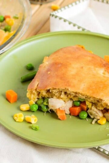 A slice of turkey pot pie on a green plate, filled with chicken, peas, carrots, corn, and a golden-brown crust, with a pie dish in the background.