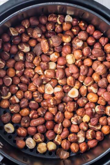 An overhead view of a large batch of air fryer hazelnuts being roasted inside an air fryer basket. The nuts are in various shades of brown, filling the circular basket to the brim.