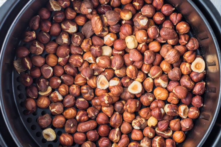 An overhead view of a large batch of air fryer hazelnuts being roasted inside an air fryer basket. The nuts are in various shades of brown, filling the circular basket to the brim.