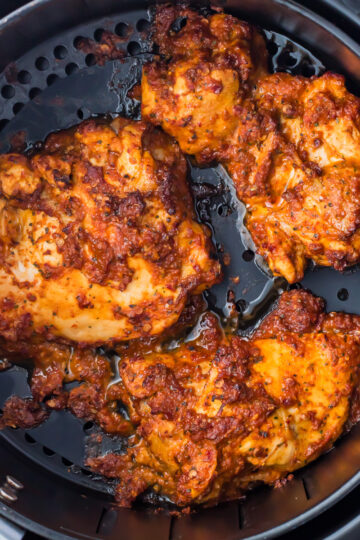 Cooked chicken pieces, seasoned with a reddish spice mix reminiscent of pollo asado, placed inside an air fryer basket. The chicken appears crispy and well-cooked.