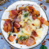 A baked camembert with bacon, basil, and a glaze of honey. The camembert is partially melted, surrounded by a sprinkle of red pepper flakes on a light blue surface.