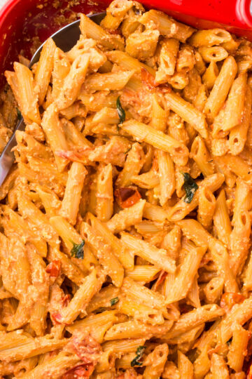 A red baking dish filled with creamy baked feta pasta mixed with cherry tomatoes and herbs. A serving spoon is placed inside the dish, ready to serve.