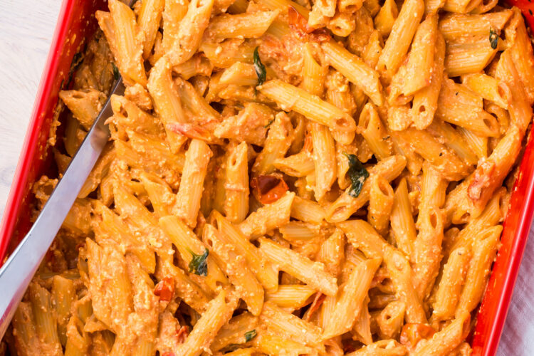 A red baking dish filled with creamy baked feta pasta mixed with cherry tomatoes and herbs. A serving spoon is placed inside the dish, ready to serve.