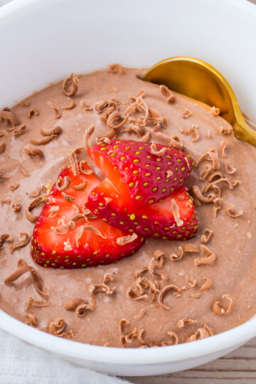 A white bowl filled with chocolate cottage cheese mousse, topped with sliced strawberries and chocolate shavings, with a gold spoon on the side.