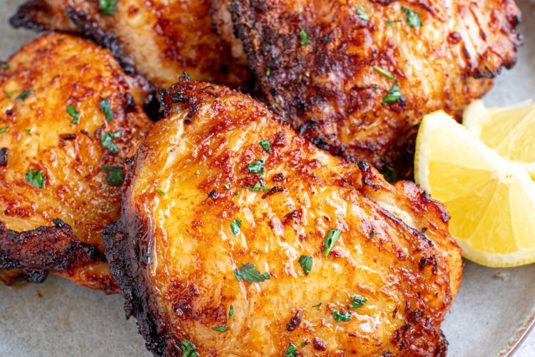 Four easy air fryer chicken thighs sit on a plate with lemon wedges on the side.