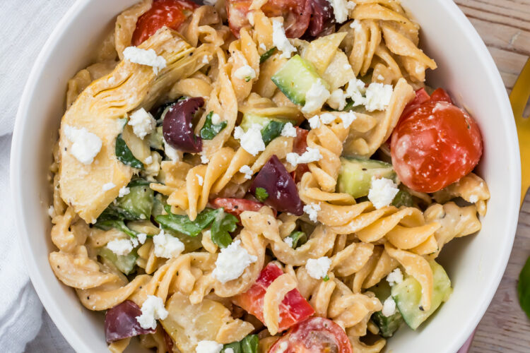 A bowl of gluten free pasta salad featuring rotini pasta, cherry tomatoes, cucumber pieces, artichoke hearts, black olives, and spinach, all topped with crumbled feta cheese. The dish is arranged on a wooden table with a white napkin nearby.