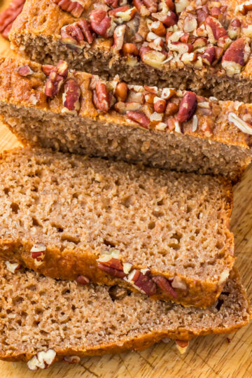 Sliced loaf of healthy banana bread topped with chopped pecans on a wooden cutting board, with a few pecans scattered nearby.