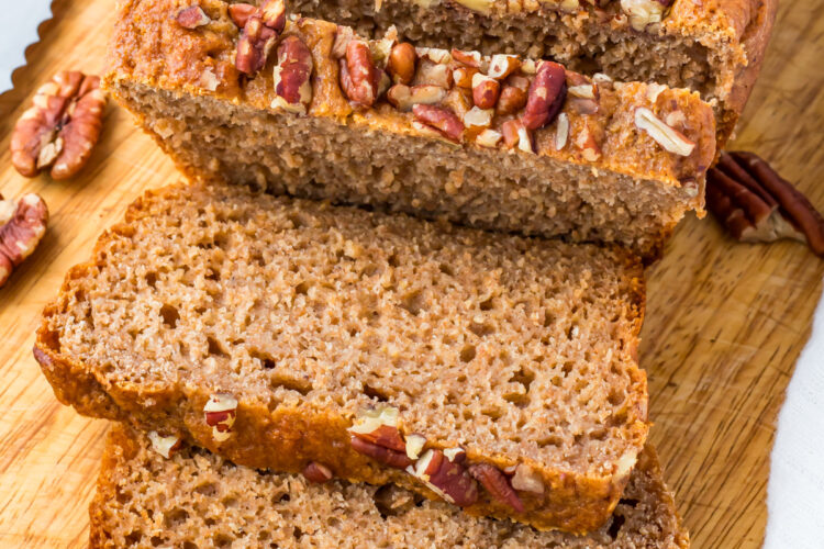 Sliced loaf of healthy banana bread topped with chopped pecans on a wooden cutting board, with a few pecans scattered nearby.