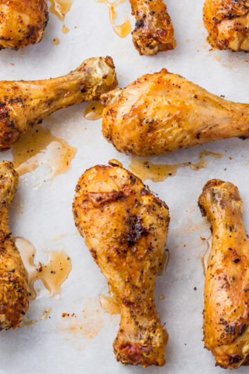 Oven baked drumsticks with a golden, crispy skin are arranged on a sheet of parchment paper. They appear well-seasoned, with visible spices and a slight glaze from their natural juices.