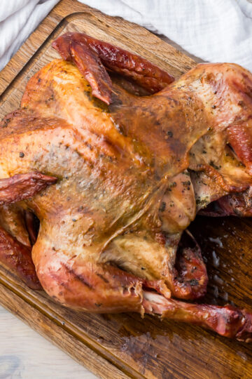 Smoked turkey with golden brown skin resting on a wooden cutting board. The turkey is seasoned and has crispy skin, with white cloth napkins partially visible in the background.
