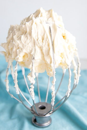 Buttercream on a whisk with a blue tea towel behind.