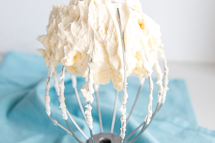 Buttercream on a whisk with a blue tea towel behind.