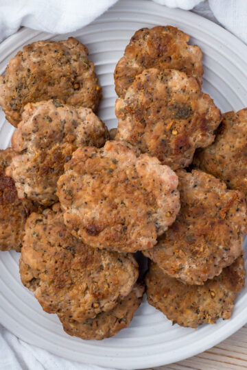 A white plate filled with several browned, seasoned turkey sausage patties, set on a light wooden surface with a white cloth in the background.