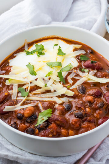 A bowl of vegan three bean chili featuring kidney, black, and pinto beans is topped with shredded cheese, sour cream, and cilantro. The bowl rests on a white cloth with a fork and a small dish of cheese nearby.