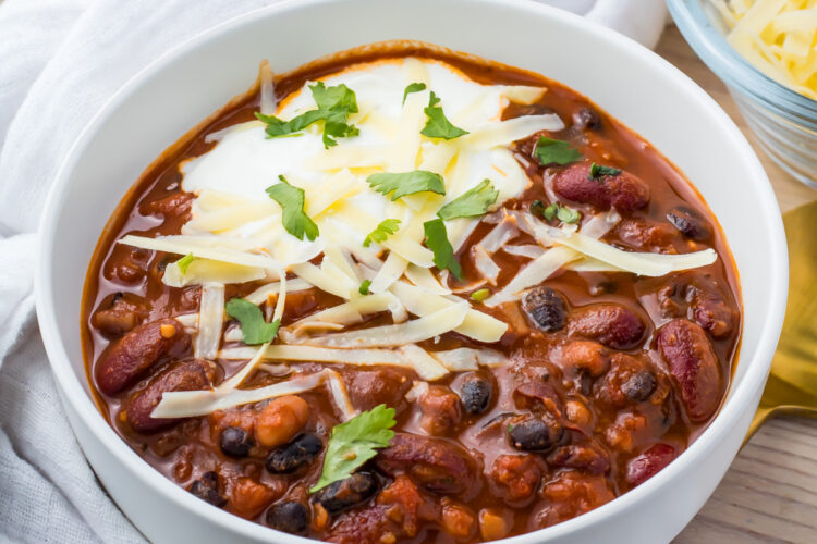 A bowl of vegan three bean chili featuring kidney, black, and pinto beans is topped with shredded cheese, sour cream, and cilantro. The bowl rests on a white cloth with a fork and a small dish of cheese nearby.