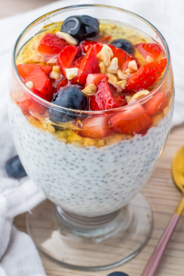 A glass of yogurt chia pudding topped with strawberries, blueberries, granola, and honey sits on a table next to a gold spoon and a white napkin.