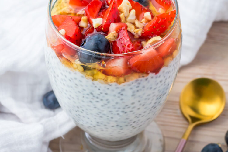 A glass of yogurt chia pudding topped with strawberries, blueberries, granola, and honey sits on a table next to a gold spoon and a white napkin.